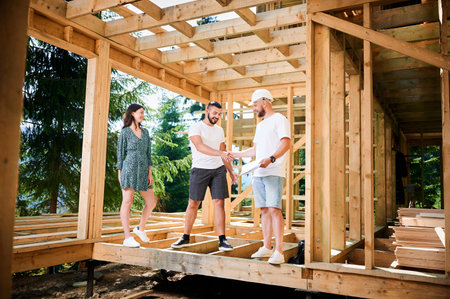 Man showcases to couple the process of constructing wooden framed house near the forest. Happy men firmly grasp each others hands in a gesture of mutual respect. Concept of reaching mutual agreement.の写真素材