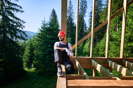 Carpenter constructing wooden frame two-story house near the forest. Bearded man holding hammer, dressed in protective helmet and overalls. Concept of ecological modern construction.の写真素材