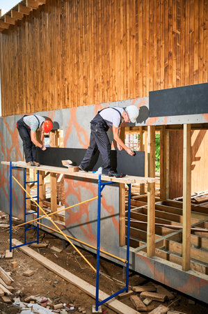 Carpenters constructing wooden framed house. Two men workers cladding facade of house with cement...