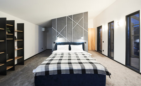 Minimalist interior of bedroom with big bed and wooden brown shelves indoor. Front view of wide bed, with plaid blanket against gray wall with geometric ornament in sleeping area. Concept of interior.の写真素材