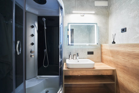 Minimalist grey bathroom with modern shower cabin and white sink on wooden countertop inside. Front view of ceramic basin and mirror with warm back lights turned on in bathroom. Concept of interior.の写真素材