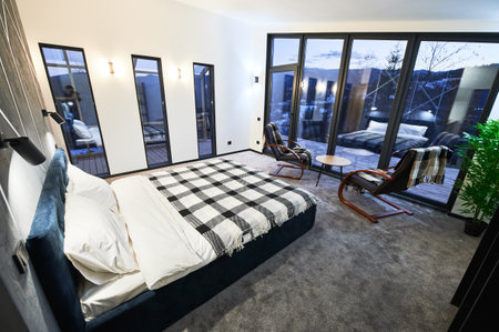Cozy modern furnished chalet room with reflection in large panoramic window at twilight. Side view of illuminated bedroom with blurred posture of photographer in mirror indoors. Concept of interior.の写真素材