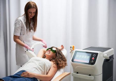 Female cosmetologist removing hair from woman underarm with diode laser device. Esthetician performing laser epilation procedure while female client in protective glasses lying on daybed.の写真素材
