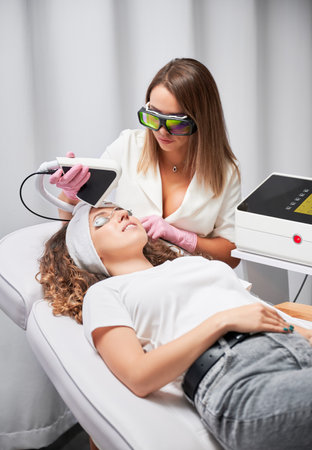 Woman patient in protective goggles receiving laser facial treatment in beauty salon. Female cosmetologist using diode laser scanner device while performing resurfacing skincare procedure.の写真素材