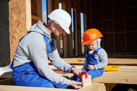 Father with toddler son building wooden frame house. Male builder learing kid to use clamp on construction site, wearing helmet and blue overalls on sunny day. Carpentry and family concept.の写真素材