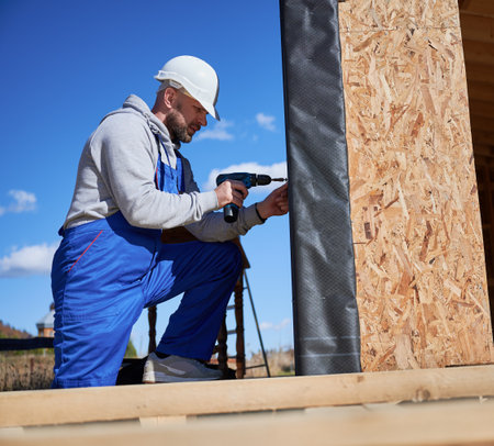 Male builder installing black corrugated iron sheet used as facade of future cottage, blue sky on background. Man worker building wooden frame house. Carpentry and construction concept.の写真素材