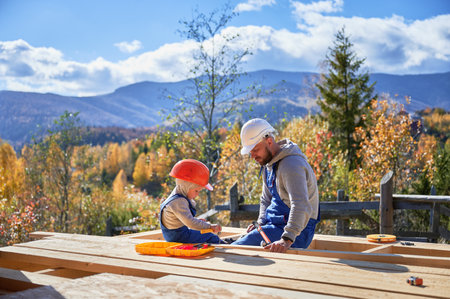 Father with toddler son building wooden frame house. Boy helping his daddy, hammering nail into plank on construction site, wearing helmet and blue overalls on sunny day. Carpentry and family concept.の写真素材