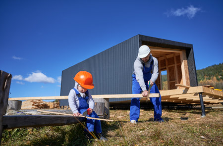 Father with toddler son building wooden frame house. Boy helping his daddy, using hand saw to cut boards on construction site, wearing helmet, blue overalls on sunny day. Carpentry and family concept.の写真素材