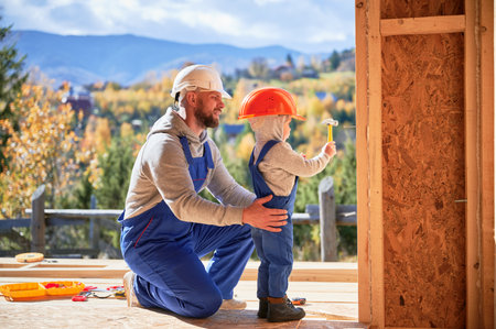 Father with toddler son building wooden frame house. Male builders hammering nail into plank on construction site, wearing helmet and blue overalls on sunny day. Carpentry and family concept.の写真素材