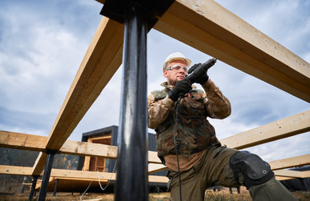 Man building wooden frame house on pile foundation. Male worker drilling hole by electric drill in timber framing. Builder wearing white helmet and protective goggles. Carpentry concept.の写真素材