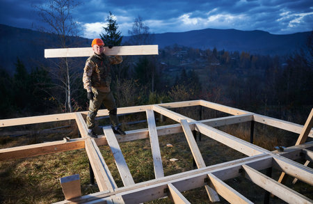 Man worker building wooden frame house on pile foundation. Carpenter carrying wooden joist for timber framing. Carpentry concept.の写真素材