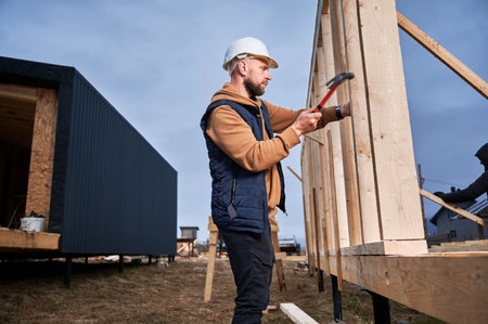 Man worker building wooden frame house on pile foundation. Carpenter hammering nail into wooden joist, using hammer. Carpentry concept.の写真素材