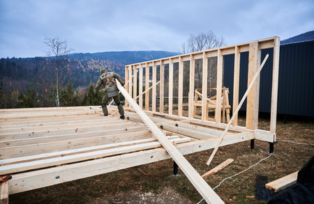 Man worker building wooden frame house on pile foundation. Carpenter carrying wooden joist. Carpentry concept.の写真素材