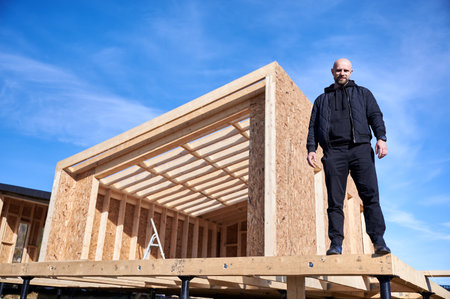 Male inspector building wooden frame house on pile foundation. Bald man standing on construction site, inspecting quality of work on sunny day with blue sky on background.の写真素材