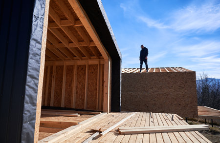 Male developer building wooden frame house. Bald man standing on construction site, inspecting quality of work on sunny day with blue sky on background.の写真素材