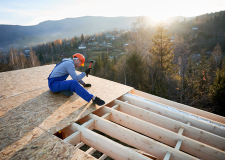 Carpenter hammering nail into OSB panel on the roof top of future cottage at sunset. Man worker building wooden frame house. Carpentry and construction concept.の写真素材
