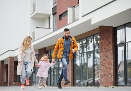 Full length of man and woman holding hands of little girl while walking down the street in new urban district. Happy loving family with child enjoying stroll outdoors near residential building.の写真素材