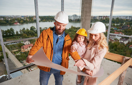 Parents with daughter studying architectural drawings in apartment building under construction. Man holding building plan and smiling while standing next to wife and daughter at construction site.の写真素材