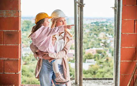Woman in building helmet holding child while standing at construction site. Kid pointing away. Mother and daughter posing in apartment building under construction.の写真素材