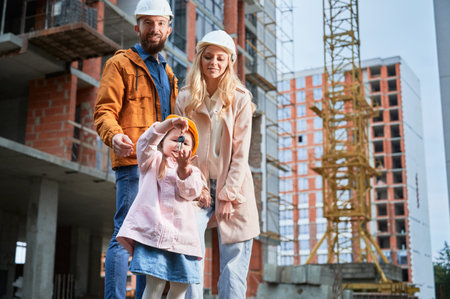 Daughter holding apartment keys and smiling while standing next to parents outside building under construction. Happy family homeowners posing on the street at construction site.の写真素材