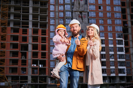 Parents and daughter showing approval gesture and smiling while standing outdoors against apartment building under construction. Happy family homeowners giving thumbs up at construction site.の写真素材