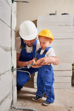 Male worker and child using wire stripper cutter tool while installing electric cables and socket in wall. Man and kid in construction helmet mounting electrical wiring in apartment under renovation.の写真素材