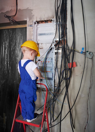 Back view of child electrician standing on ladder near electrical switchboard in apartment under renovation. Kid in safety helmet and work overalls repairing electric control panel at home.の写真素材