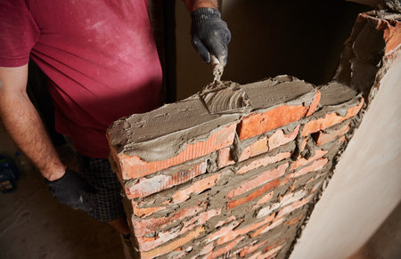 Close up of male hands in work gloves laying brickwork in building under construction. Man bricklayer builder applying cement mortar on bricks with trowel tool. Masonry construction concept.の写真素材