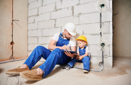 Male builder sitting by brick wall and pointing at clipboard while showing documents to little boy. Man and kid wearing safety helmets and work overalls while working on home renovation.の写真素材