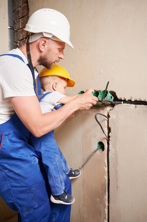 Male electrician holding adorable toddler boy while child installing electrical sockets in wall. Man and kid in safety helmet mounting electrical wiring in apartment under renovation.の写真素材