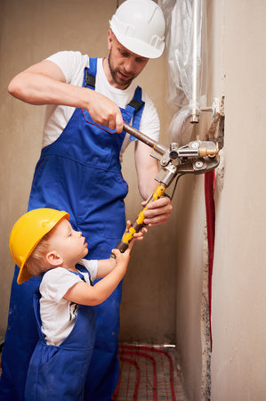 Man and child using pipe pressing tool while installing plumbing pipes in bathroom. Male plumber and kid in safety helmets working on sanitary equipment installation in apartment under renovation.の写真素材