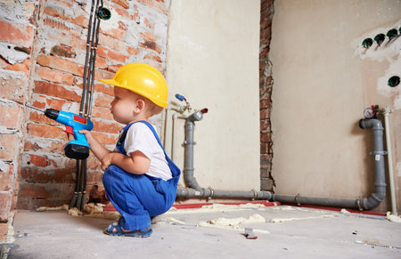 Kid construction worker crouching down and drilling brick wall with cordless electric drill. Baby boy in safety helmet using drilling power tool while working on home renovation.の写真素材