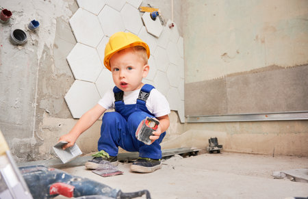 Adorable baby boy construction worker crouching down and looking at camera while holding tape measure. Cute kid wearing safety helmet and work overalls while working on home renovation.の写真素材