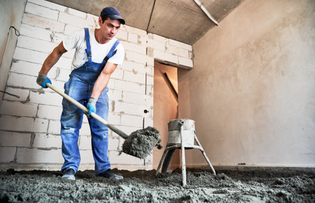 Man preparing floor screed material while standing near concrete screed mixer machine. Male worker using shovel while shoveling sand-cement mix in building under construction.の写真素材
