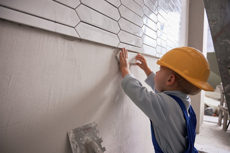 Little boy construction worker installing ceramic tile on the wall in apartment under renovation. Kid wearing safety helmet and work overalls while working on bathroom renovation.の写真素材