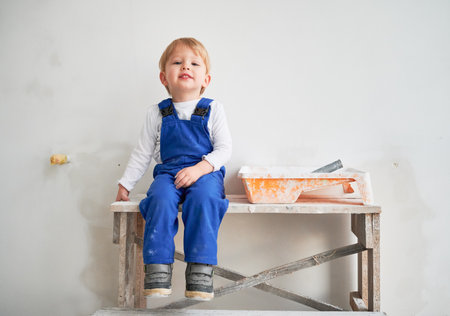 Cute kid construction worker sitting on wooden table against white wall in apartment under renovation. Cheerful little boy wearing safety helmet and work overalls while playing at home.の写真素材