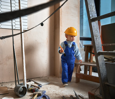 Boy construction worker with brush in apartment under renovation. Child wearing safety helmet and work overalls while playing at home.の写真素材