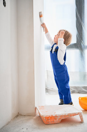 Full length of child construction worker painting wall with paint brush in apartment. Kid in work overalls using paint brush while playing at home under renovation.の写真素材