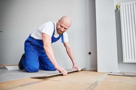 Male construction worker laying laminate wooden plank on floor underlayment. Man in work overalls kneeling down by the wall and installing laminate flooring in living room. Home renovation concept.の写真素材