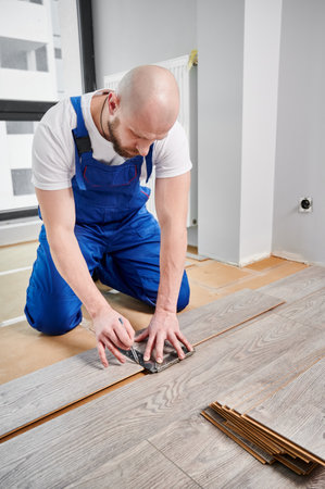 Man in work overalls using metal construction ruler and pen while drawing line on laminate flooring board. Male floor installer preparing laminate plank for installation in apartment under renovation.の写真素材