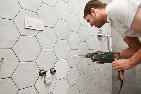 Side view of male worker drilling wall with cordless power drill in apartment under renovation. Man using electric drilling machine while working on bathroom renovation and installing shower system.の写真素材