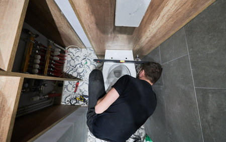 Top view of man installing flush toilet bowl in apartment under renovation. Male plumber crouching down on the floor with construction tools while working on toilet installation at home.の写真素材