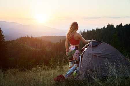 Woman tourist camping outdoors at sunset. Back view of slim woman near tent, admiring landscape in mountains. Female tourist wearing sportswear traveling in summer. Concept of beauty of nature.の写真素材
