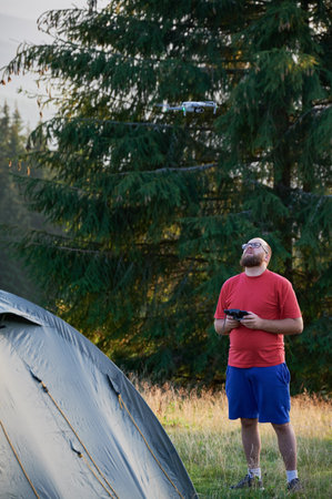 Man launching drone while camping outdoors. Young male with beard wearing glasses, standing in campsite near tent. Concept of tourism and adventure.の写真素材