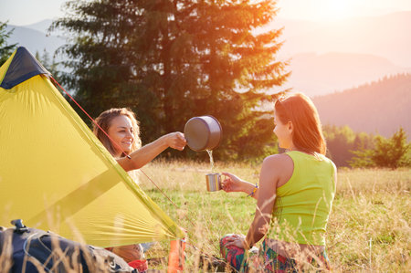 Two women travellers camping outdoors. Young women sitting in campsite, having break. Friends sitting on grass, talking, smiling, pouring water, making tea. Concept of tourism and hiking in mountains.の写真素材