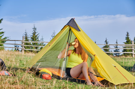 Woman hiker camping in mountains. Female tourist hiking outdoors, sitting in tent. Beautiful, young woman having break, drinking tea. Concept of beauty of nature.の写真素材