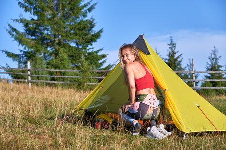 Woman traveller camping in mountains. Back view of slim, young female traveling, hiking in hills. Blonde woman with curly hair sitting on grass, looking at camera. Concept of harmony with nature.の写真素材