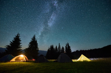 Night camping in mountains under starry sky. Tourist tents in campsite near burning campfire under beautiful sky full of stars with Milky way above forest. Concept of tourism and traveling.の写真素材
