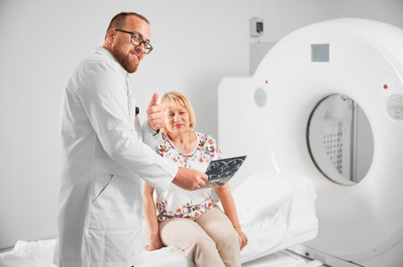 Medical computed tomography or MRI scanner. Doctor holding results of MRI and showing thumbs up. Woman patient after MRI, sitting on CT scan bed. Concept of medicine, healthcare, modern diagnostics.の写真素材