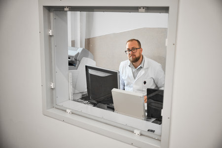 Doctor sitting, working at computer, making MRI. Male specialist wearing glasses and white robe, looking at camera, smiling, Concept of modern diagnostics.の写真素材